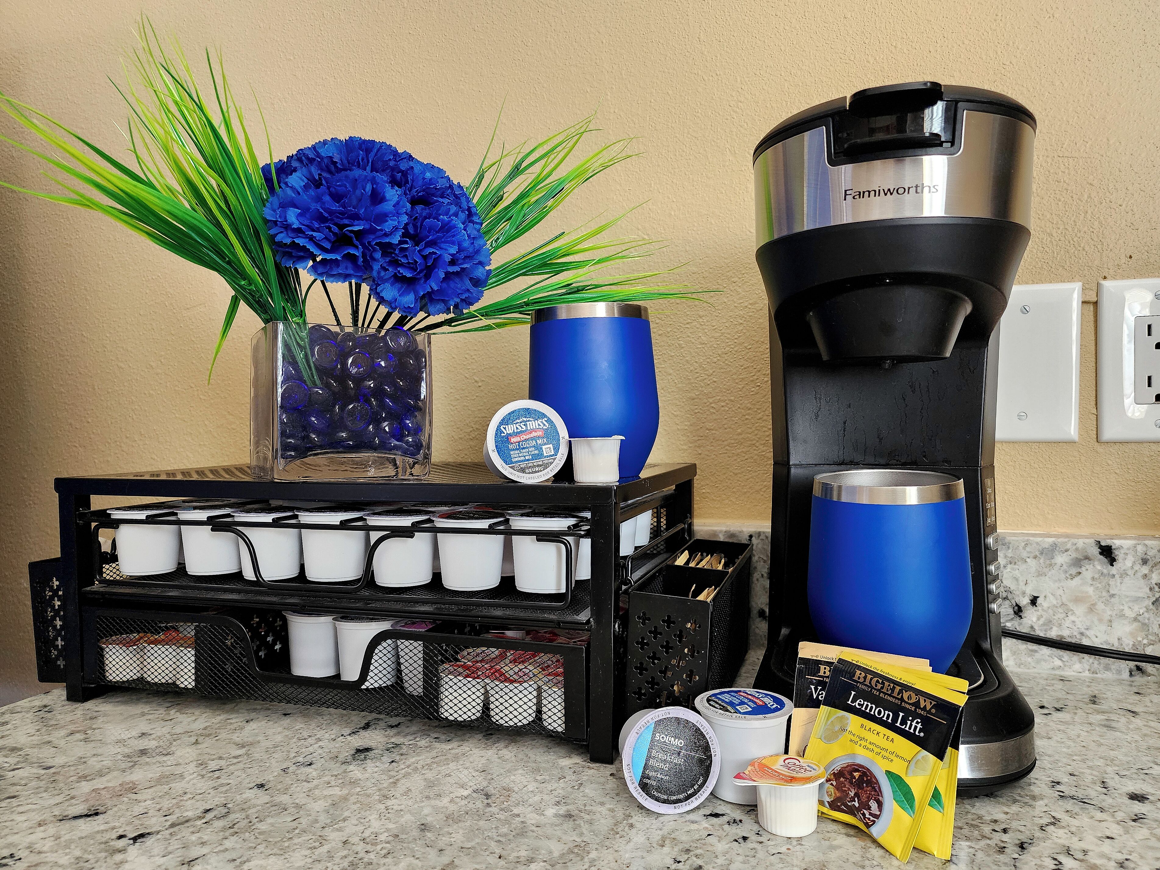 A kitchen counter with a coffee maker, a tray of coffee cups, a vase with blue flowers, and tea bags.