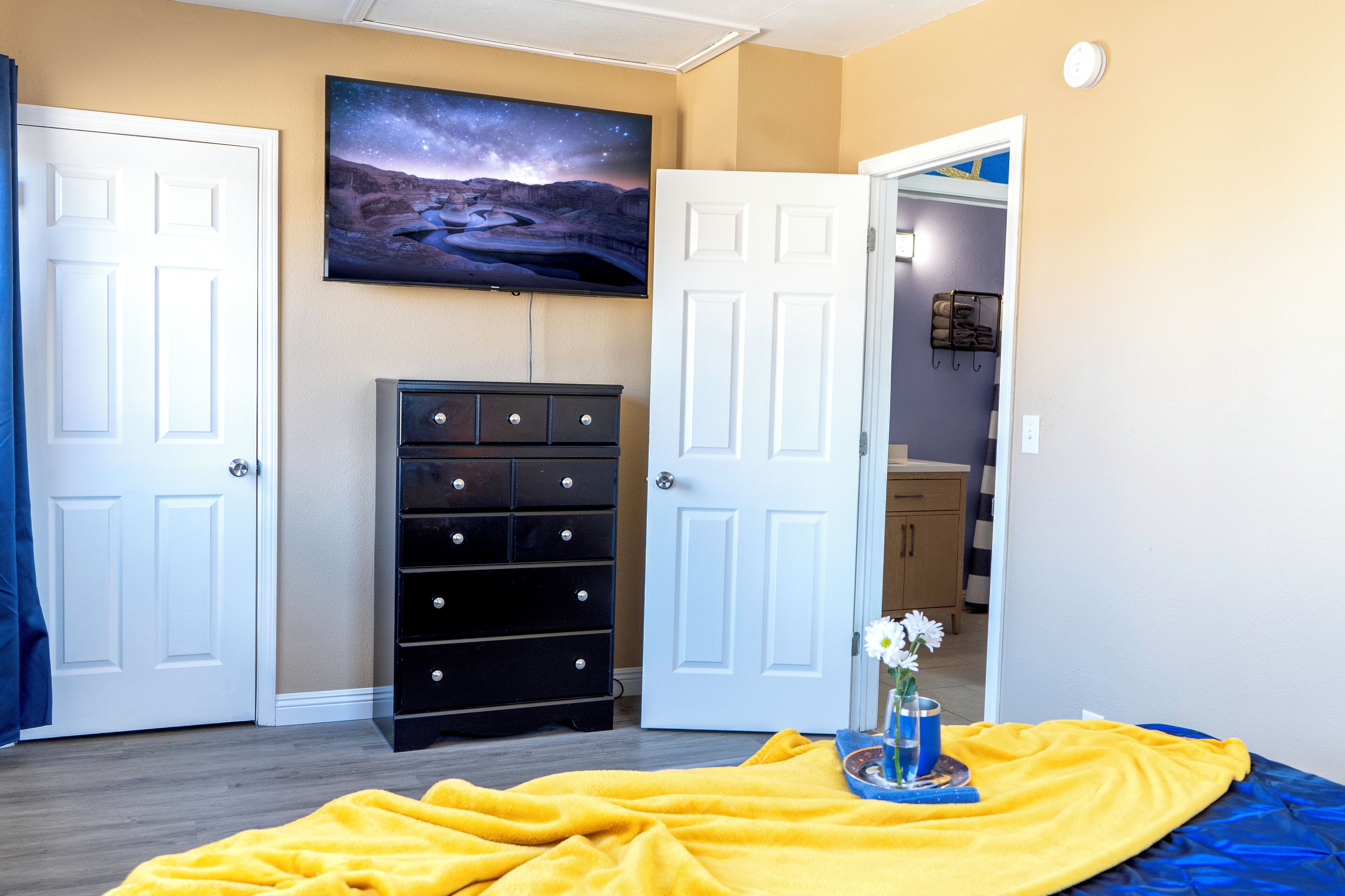 Ein Schlafzimmer mit zwei weißen Türen, einer schwarzen Kommode, einem Bett mit gelber Decke, einem Bild von Sternennacht an der Wand und einer kleinen Vase mit Blumen auf dem Bett.