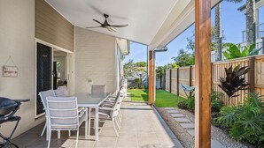 Un patio al aire libre con una mesa, sillas, un ventilador de techo y vistas a un jardín y casas vecinas.