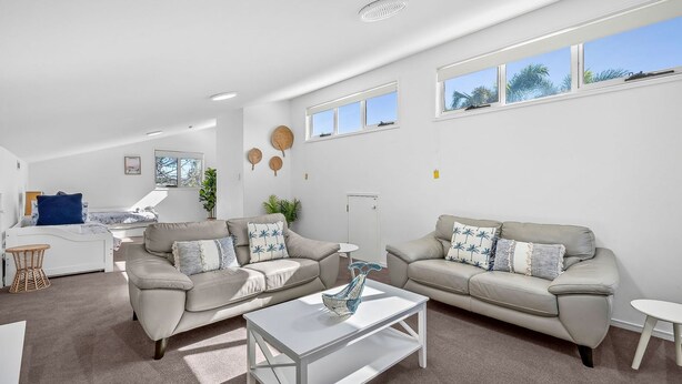 Una sala de estar moderna con dos sofás color beige, una mesa de centro blanca y una pequeña mesita auxiliar blanca. Hay dos ventanas que proporcionan luz natural, y se ve una planta en la esquina.