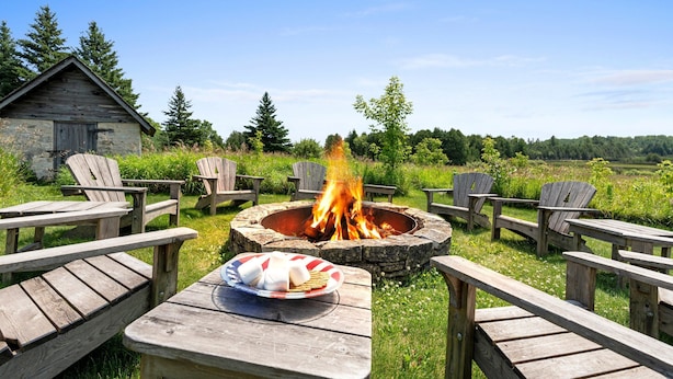 Un cadre extérieur rustique avec un foyer en pierre entouré de fauteuils Adirondack en bois, une table en bois sur laquelle se trouve une assiette de nourriture, et un chalet en bois en arrière-plan.