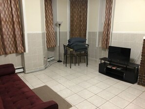 Un salon comprenant un canapé, un meuble pour la télévision avec un téléviseur à écran plat, une table avec des chaises, des rideaux à motifs et un revêtement de sol carrelé.
