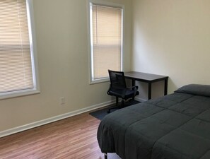 A bedroom with a bed, a desk, and two windows with blinds.