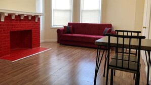 Una sala de estar con chimenea de ladrillo rojo, un sofá rojo, una mesa de comedor de madera y dos ventanas con persianas.