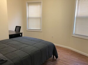 A bedroom with a bed, a desk, and two windows with blinds.