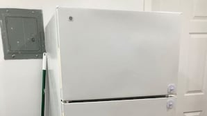 Een witte koelkast in een kamer met houten vloeren en een bezem die tegen de muur staat.