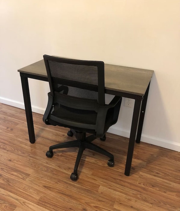 Una silla de oficina negra con respaldo de malla y ruedas, ubicada sobre un piso de madera junto a un escritorio con acabado oscuro.