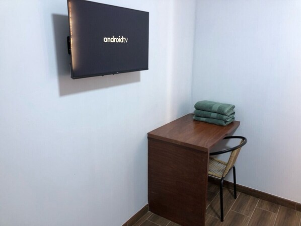 A small room with a wooden desk, a chair, a flat-screen TV mounted on the wall, and a stack of folded towels on the desk.