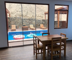 A room with a wooden table and chairs, a view of a swimming pool with lounge chairs and inflatable rings, and a stone wall outside.