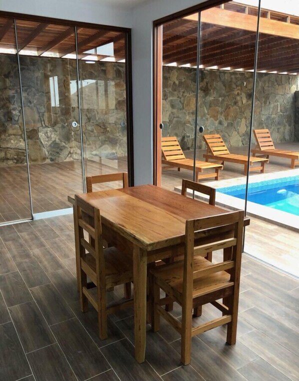 Une table à manger en bois, avec des chaises, dans une pièce dont un mur est en pierre et qui possède une porte en verre donnant sur un espace piscine.