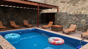 A rectangular pool with two inflatable rings, surrounded by wooden lounge chairs and a stone wall. - Spacious 3-bedroom and 2 bathroom home with private pool and patio and free Wi-Fi (Calango)