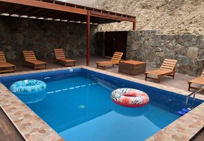Une piscine rectangulaire avec deux bouées gonflables, entourée de chaises longues en bois et d’un mur en pierre.