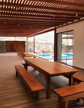 A covered outdoor seating area with wooden benches and tables, a stone wall, and a view of a swimming pool.