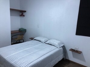 Une petite chambre à coucher, bien rangée, avec un lit simple, une chaise, une étagère sur laquelle est posée une serviette verte et un téléviseur fixé au mur.