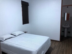 A bedroom with a bed, pillows, a nightstand, a window with blinds, and a bathroom with a sink and mirror.