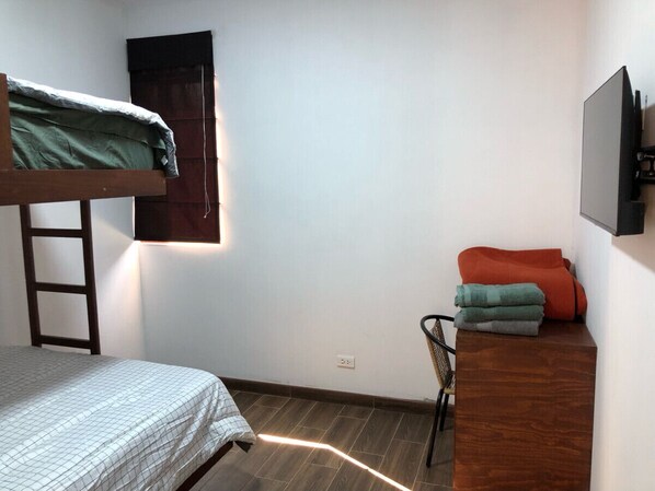 Une petite pièce, peu meublée, avec un lit superposé, un bureau avec une chaise, un petit meuble contenant des serviettes et un téléviseur fixé au mur.