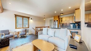 A modern living room with a sofa, chairs, a small table, and a kitchen area with wooden cabinets and a refrigerator.