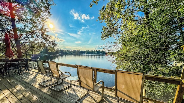 Terraza de madera con sillas que da a un lago tranquilo, rodeada de árboles y un cielo despejado.
