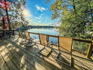 A wooden deck with chairs overlooking a calm lake, surrounded by trees and a clear sky.