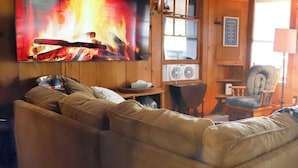 Interior acogedor de una cabaña con un amplio sofá modular, una chimenea y un televisor montado encima, además de un letrero que indica la dirección hacia 'LAKE'.