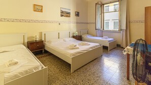 Habitación de hotel con dos camas, una mesita de noche con lámpara, una ventana con contraventanas y un ventilador.