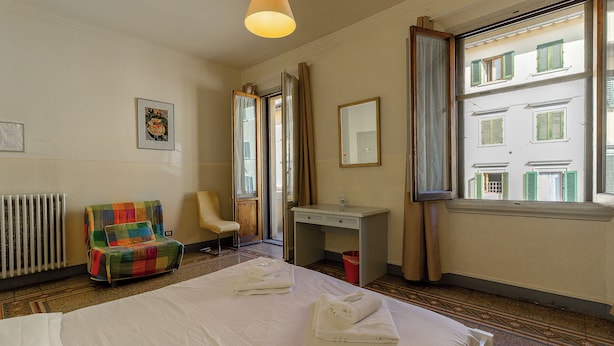 Una habitación de hotel con cama, un sillón colorido, un escritorio con espejo y una ventana con contraventanas.