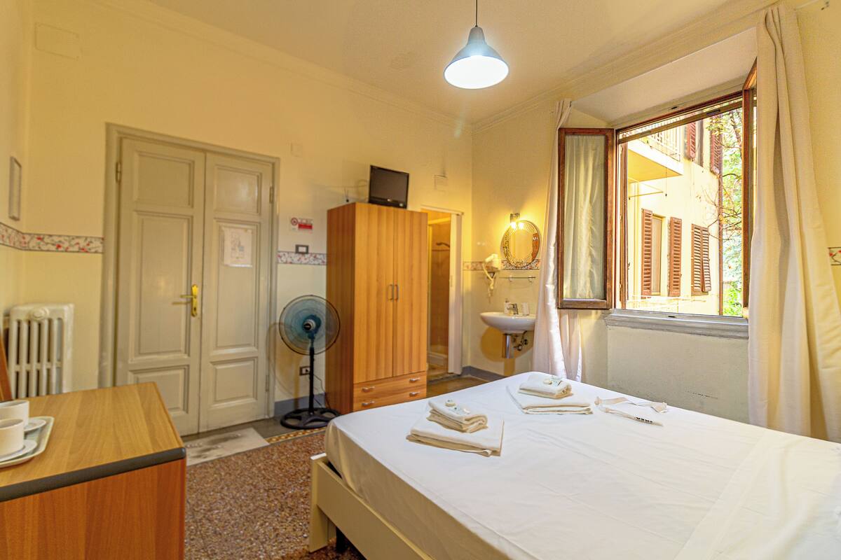 Habitación de hotel con cama, armario de madera, escritorio con taza, ventilador, ventana con cortinas y baño con lavamanos y espejo.
