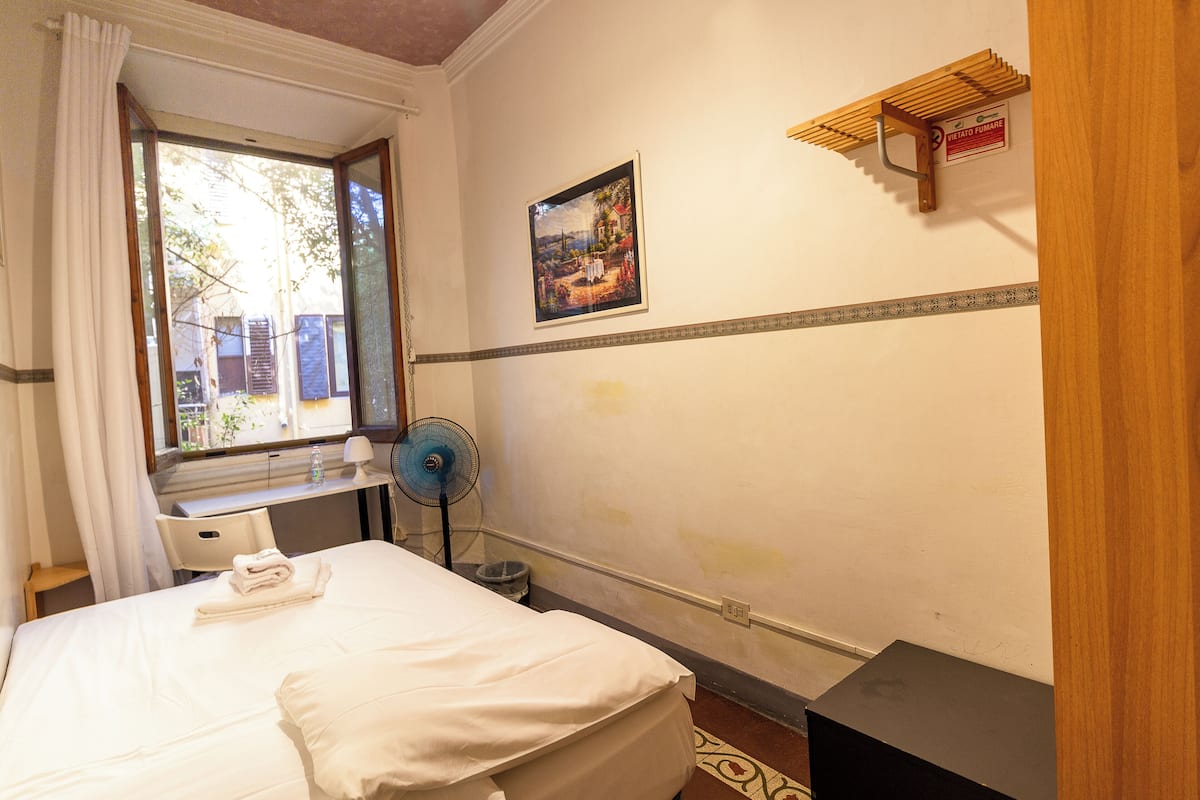 Una habitación de hotel con una cama, una ventana con cortinas, un escritorio con una silla, un ventilador y una pequeña estantería con un cartel.
