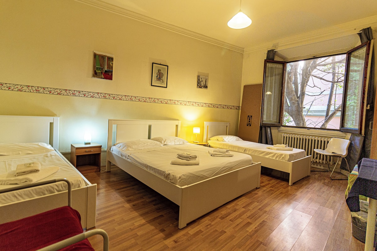 Habitación de hotel con dos camas individuales, una mesita de noche con lámpara, una silla, un radiador y una ventana con vistas a árboles.