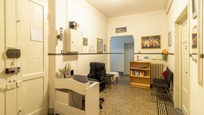 Un consultorio clínico con una cama, un escritorio con computadora, una silla, una estantería y un radiador.