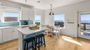 A modern kitchen with white cabinetry, a large island with stools, a view of the ocean, and a balcony with a wooden deck.