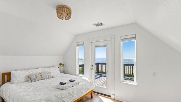 A bedroom with a large bed, a wooden floor, and a view of the beach through a door.
