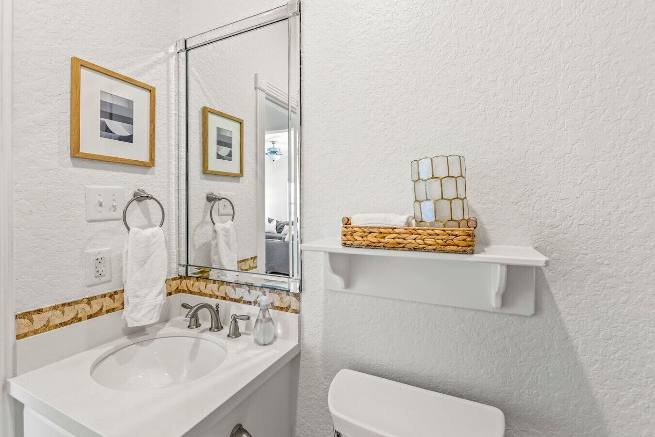 A bathroom with a white sink, two faucets, a mirror, and a toilet. There are two framed pictures on the wall, a towel ring, and a woven basket on a shelf.