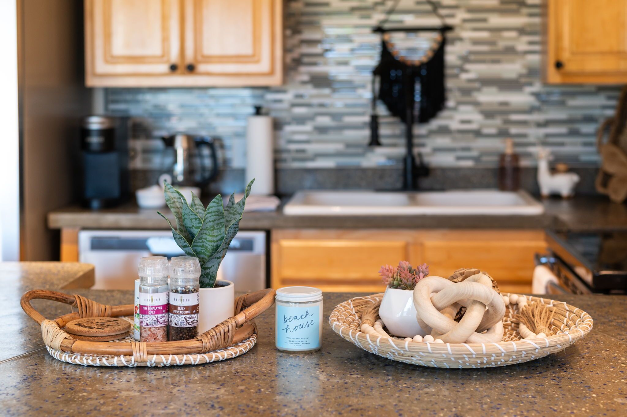 A kitchen counter with a coffee maker, a plant, a jar labeled 'beach house', and a basket with pretzels and a woven item.
