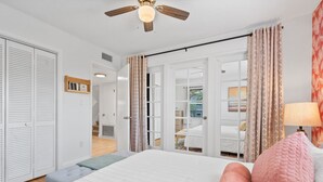 A bedroom with a bed, a ceiling fan, a bedside lamp, and a view of another room through an open door.