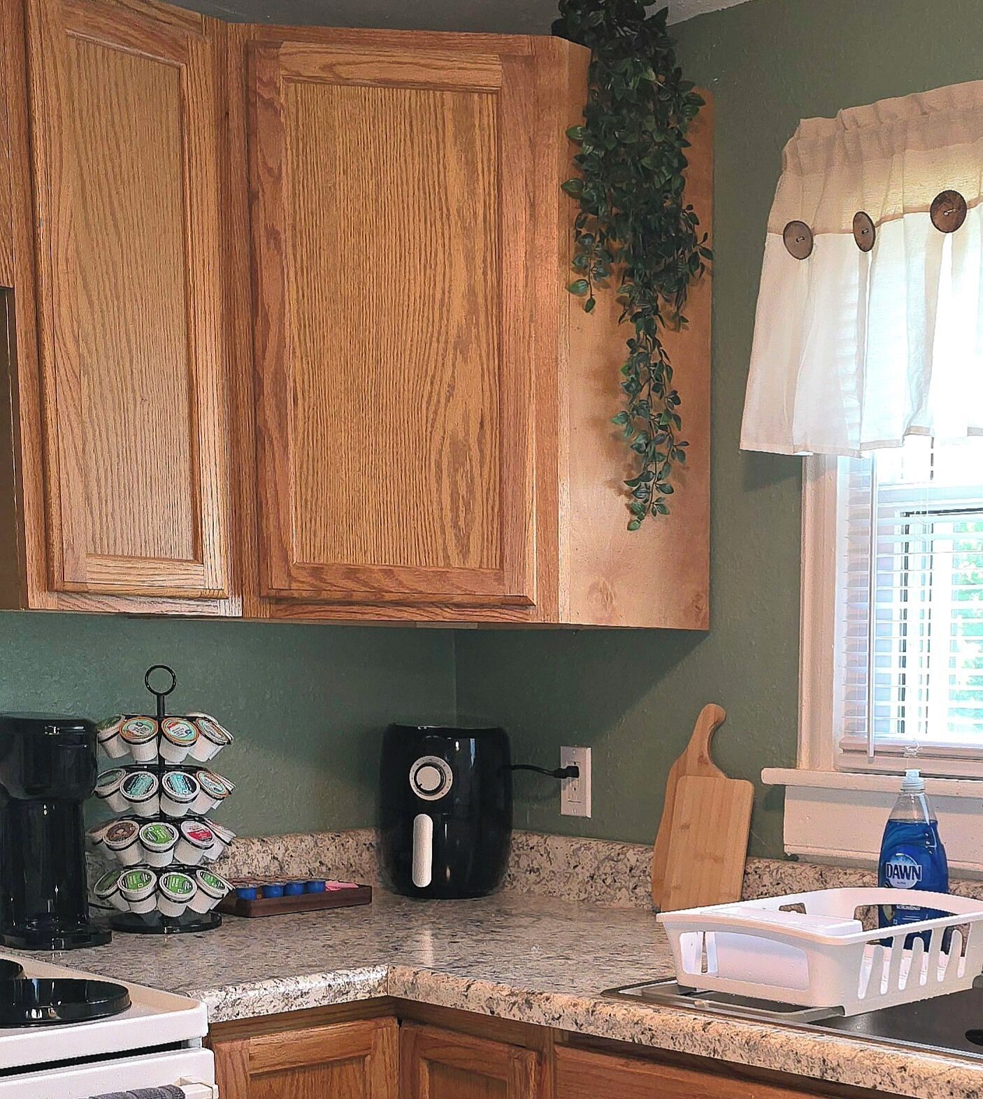 A kitchen with wooden cabinets, a coffee maker, a plant, a cutting board, and a window with curtains.