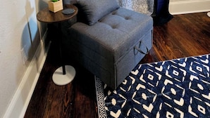 Una sala de estar con un sofá gris, un tapete estampado, una pequeña mesita de madera y una ventana con cortinas azules.