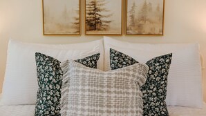 A bed with white pillows and floral-patterned pillows, framed artwork above, and a plain wall.