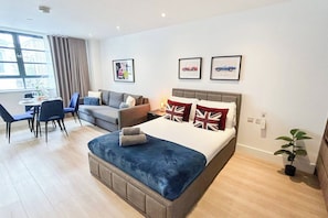 A modern hotel room with a bed, a sofa, a dining table with chairs, and a window with curtains.