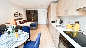 Un espacio habitable compacto con comedor, kitchenette y una pequeña zona de estar con estantería.