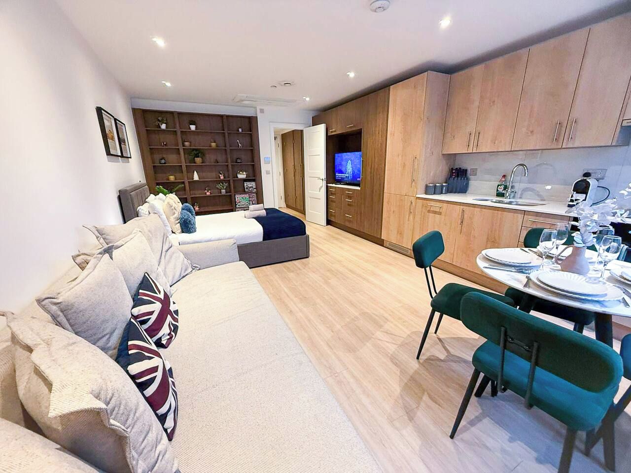 A modern living room with a sofa, a dining area with a table set for two, and a kitchen with wooden cabinets and a TV.