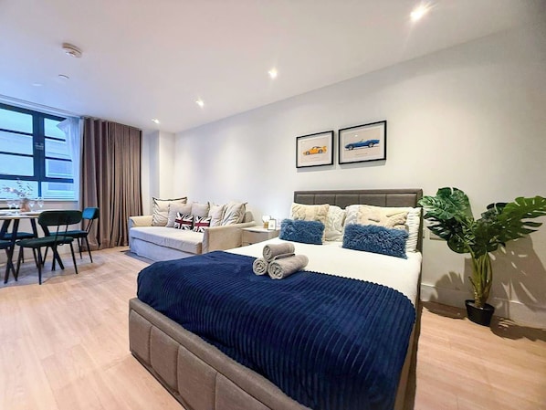A modern hotel room with a large bed, a sofa, a dining area, and a potted plant.