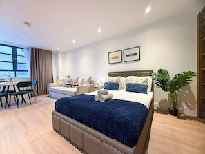 A modern hotel room with a large bed, a dining area, and a view of the city.