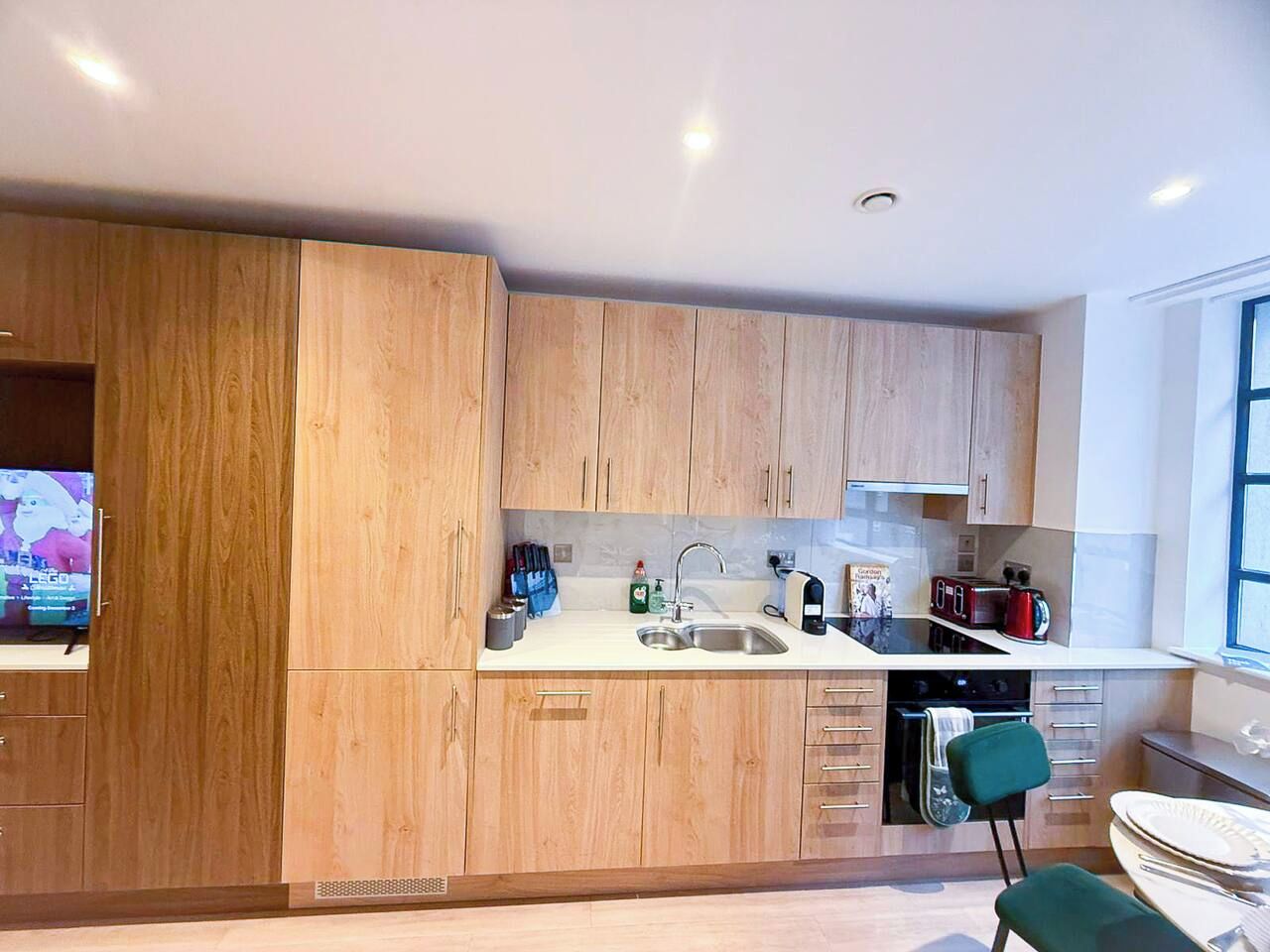 A modern kitchen with wooden cabinets, a sink, a stove, and a dining area with a green chair.