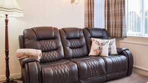 Un sofá de cuero marrón con diseños acolchados, un cojín decorativo, una lámpara de pie y cortinas a cuadros.