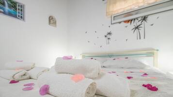 A neatly made bed with rolled white towels and pink heart-shaped bath items, a wooden headboard, and a wall with palm tree and bird decals.