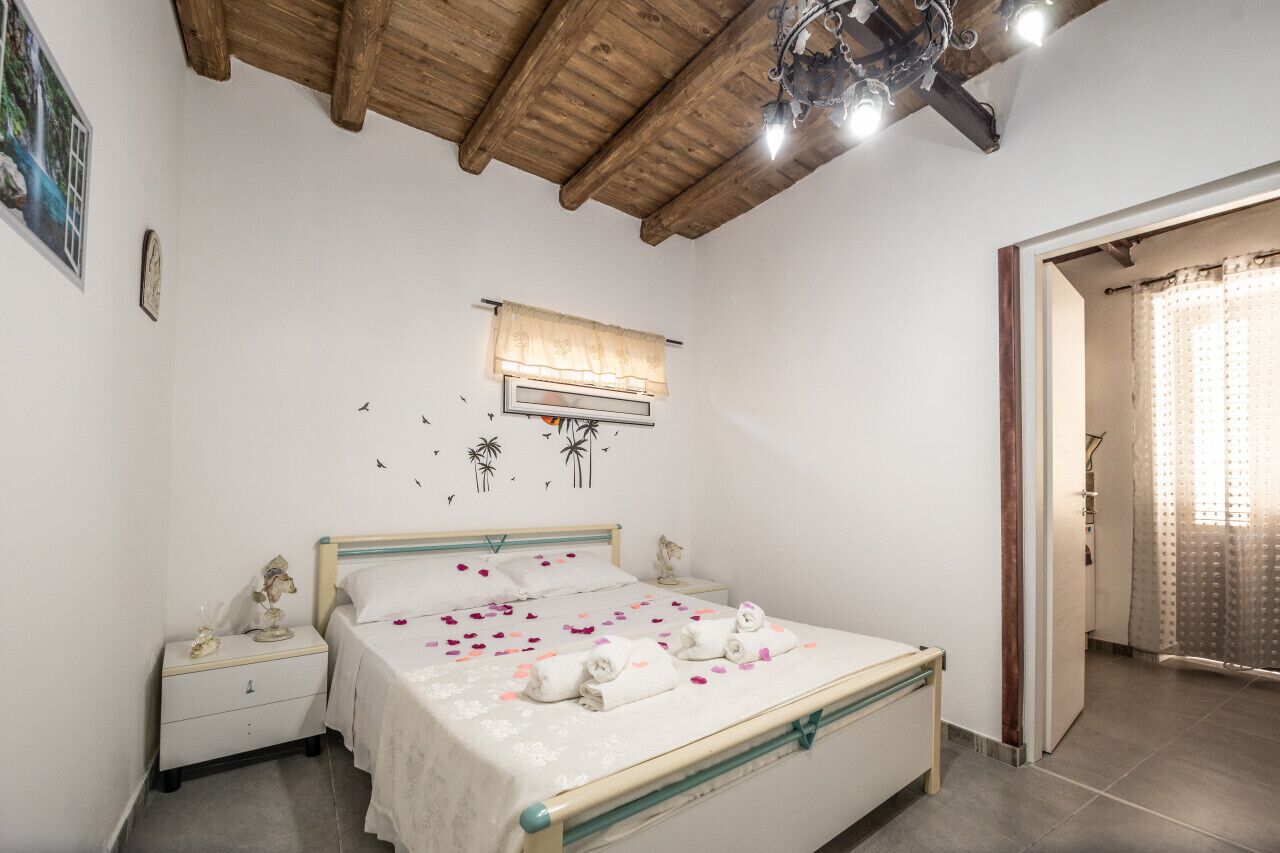 A bedroom with a wooden ceiling, a bed with white linens, a nightstand, a wall-mounted air conditioning unit, and a door with a curtain.