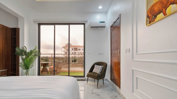 Une chambre moderne avec un grand lit, une porte-fenêtre coulissante donnant sur un balcon, une plante en pot et un climatiseur fixé au mur.