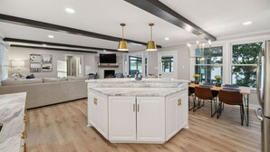 A modern kitchen with a central island, white cabinetry, and gold handles. There is a dining area with a wooden table and chairs.