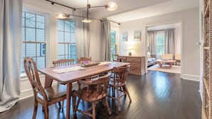 Un comedor con mesa y sillas de madera, una araña y ventanas grandes con cortinas.